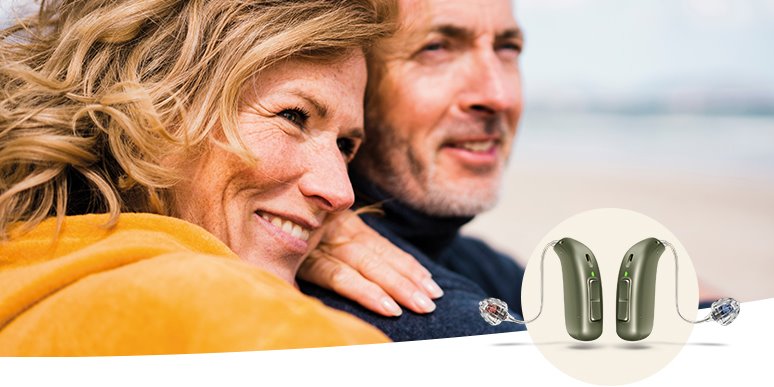 Dos audífonos modernos de color gris con diseño discreto se muestran destacados sobre un círculo blanco. Al fondo, una escena difuminada con personas abrazándose y un entorno costero.