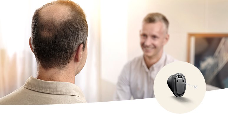 Un hombre de espaldas consulta a un audioprotesista en una clínica auditiva iluminada; se destaca un audífono intracanal negro moderno visto en detalle a la derecha.