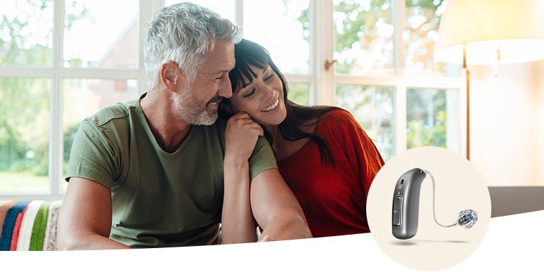 Una pareja sentada en un ambiente hogareño con grandes ventanas al fondo. A la derecha, un audífono discreto de tecnología avanzada, destacado sobre un fondo blanco.