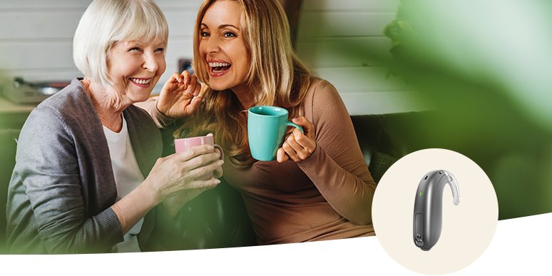 Dos mujeres sentadas en un sofá, compartiendo una conversación amigable mientras sostienen tazas de té o café; en la esquina, se muestra un audífono retroauricular Audika sobre un fondo blanco.