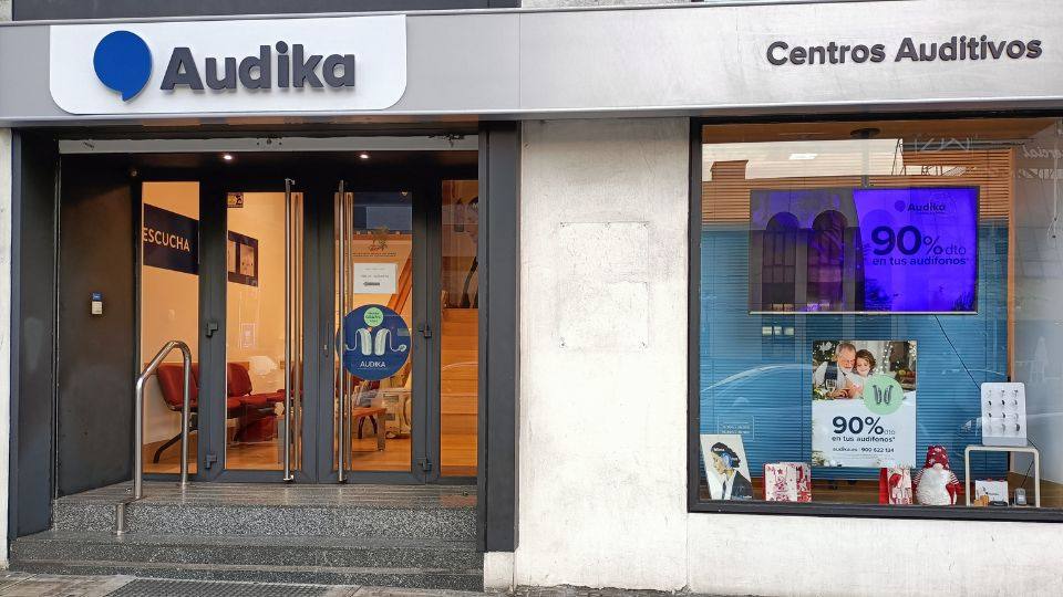 La fachada de un centro auditivo Audika muestra una entrada con puertas de vidrio y un cartel que dice 'ESCUCHA'; junto a una vitrina promocional con el texto: '90% dto en tus audífonos'.