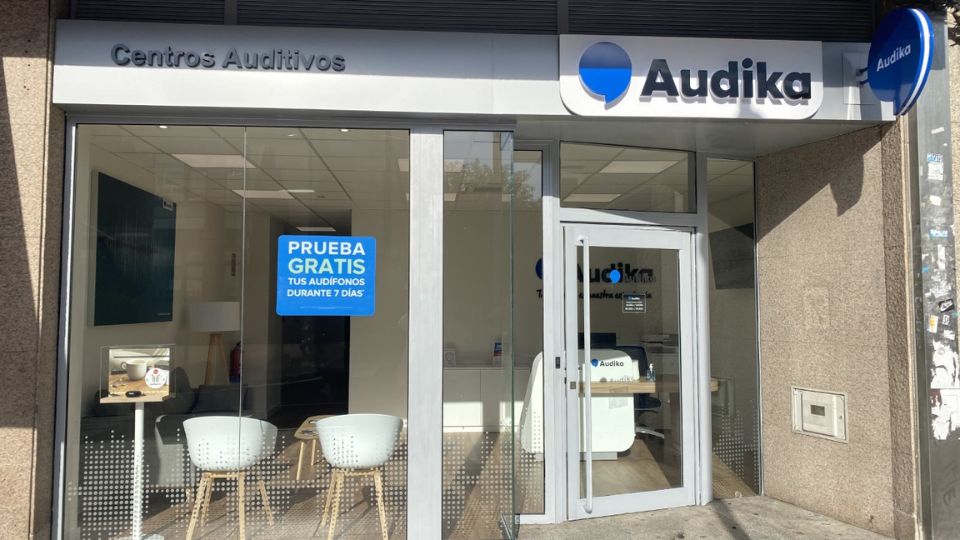 Fachada de un centro auditivo Audika con cristaleras, mostrando su interior moderno con sillas blancas. Un cartel dice: ’PRUEBA GRATIS TUS AUDÍFONOS DURANTE 7 DÍAS’.