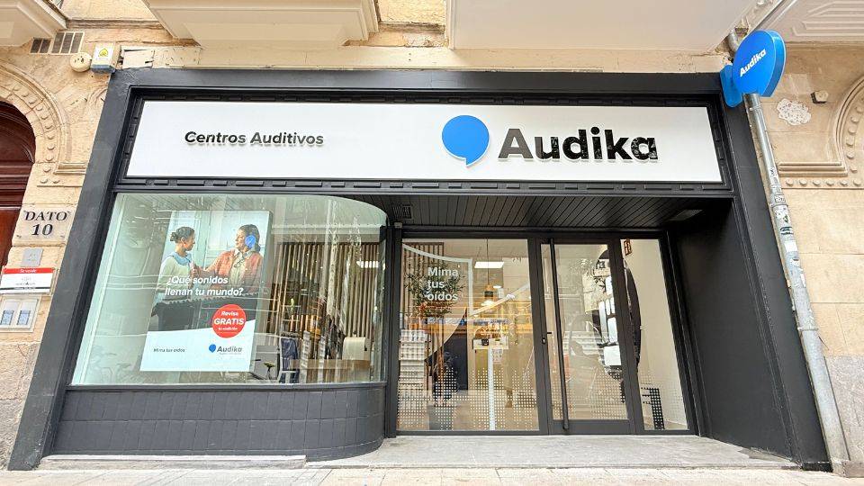 Fachada de un centro auditivo Audika con letrero 'Centros Auditivos'. Un cartel muestra: '¿Qué sonidos llenan tu mundo? Prueba GRATIS. Visita tus expertos Audika'. Contexto urbano.