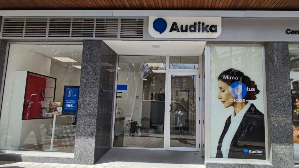 Fachada de una clínica auditiva Audika, con puertas de vidrio y carteles promocionales. Texto visible: ’Mima tus oídos’ y ’Tu audífono desde 49€’. Interior luminoso con diseño moderno.