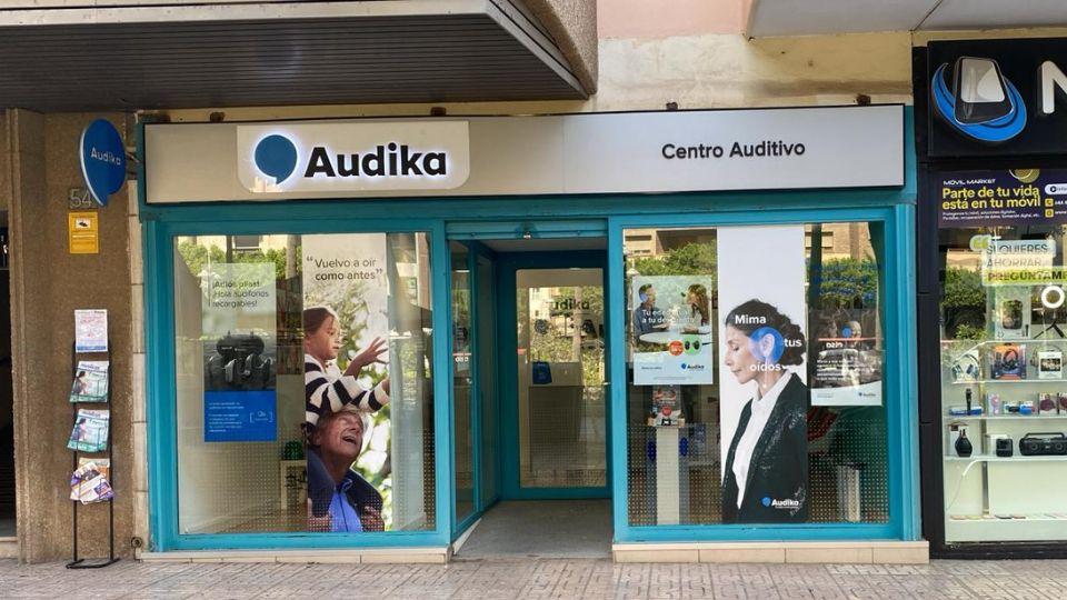 Fachada de un centro auditivo Audika con ventanas de cristal decoradas con carteles promocionales: “Vuelve a oír como antes' y “Mima tus oídos'. Ubicación en entorno comercial urbano.