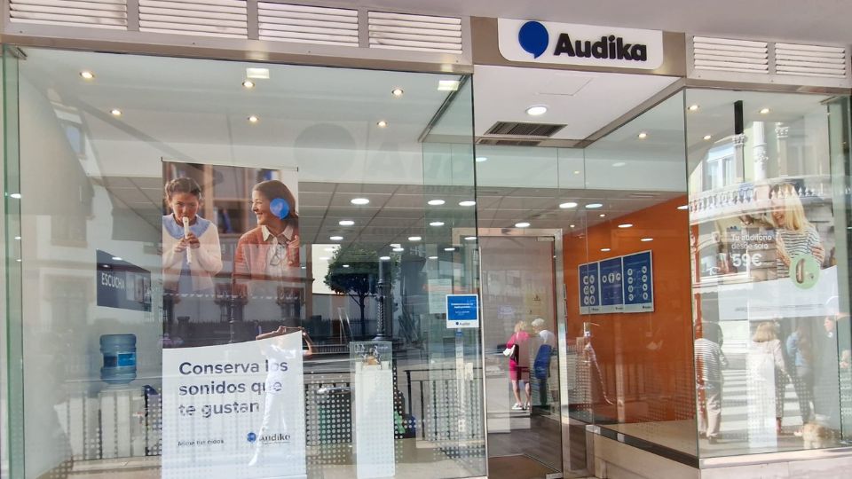 Fachada de una clínica auditiva Audika con puerta de vidrio; carteles promocionales destacan: **'Conserva los sonidos que te gustan'**, precios desde 59€, y atención especializada en audífonos. Ambiente ordenado y moderno.