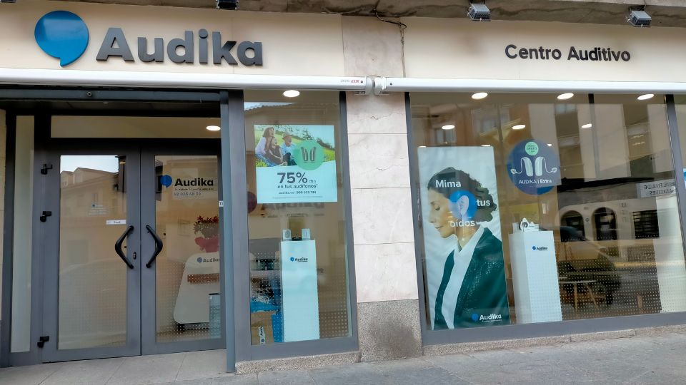 Fachada de un centro auditivo de Audika con ventanas de cristal, el logo visible en la parte superior izquierda y carteles promocionales como ’75% en tus audífonos’ y ’Mima tus oídos’.