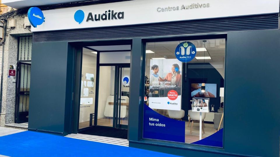 La fachada de un centro auditivo Audika muestra un diseño moderno con ventanales. En el cristal destacan textos: ’¿Qué sonidos llenan tu mundo?’ y ’Revisión GRATIS’. Ambiente acogedor.