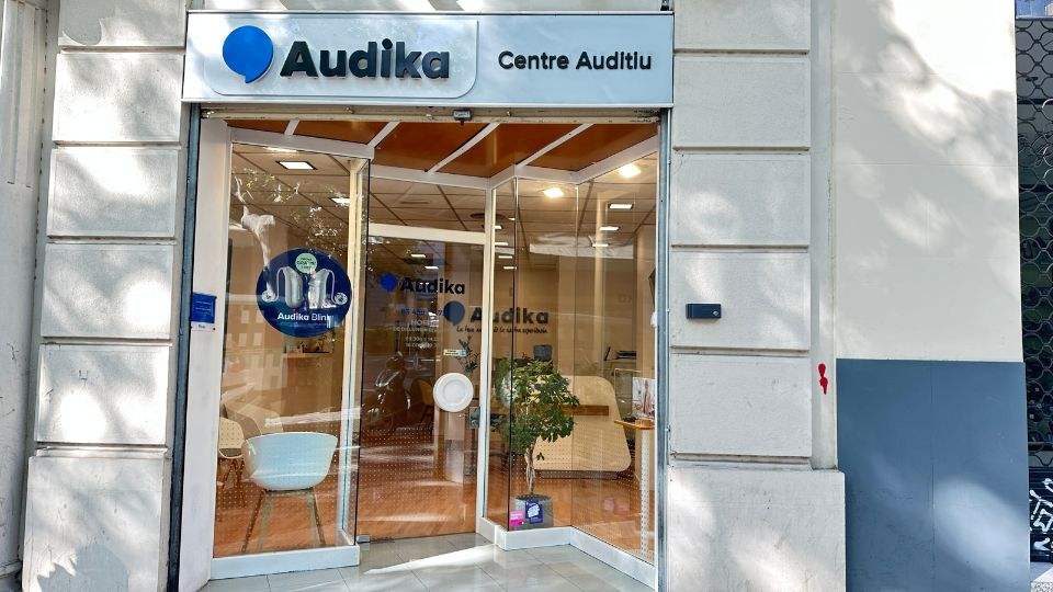 La fachada de un centro auditivo Audika, con puertas de vidrio que muestran el interior iluminado, plantas decorativas y sillas blancas. Cartel: 'Audika Direct' y 'Centre Auditiu'.