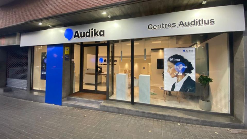 Fachada de un centro auditivo Audika con rótulo 'Centres Auditius'. Interior bien iluminado, con áreas visibles para prestaciones como audífonos y pruebas auditivas. Cartel promocional: 'Mima la teva oïda'.