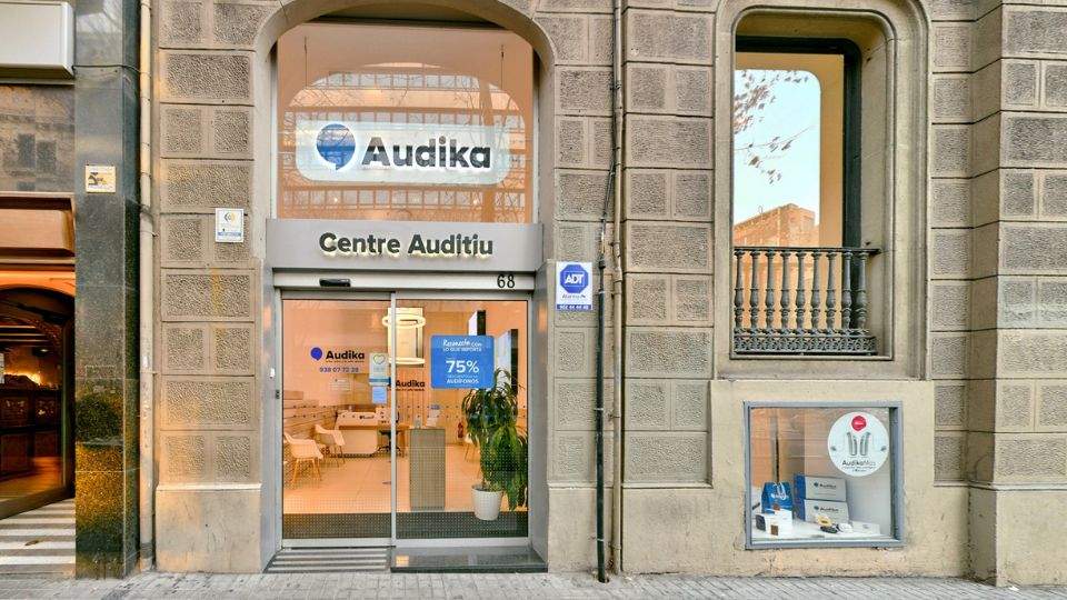 Fachada de Audika, Centre Auditiu, en el número 68. Puerta de vidrio con carteles promocionando descuentos del 75% en audífonos. Escaparate lateral exhibe audífonos y material de Audika.