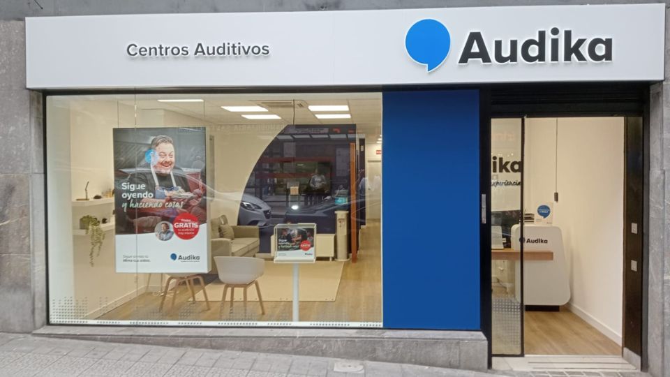 Fachada de un centro auditivo Audika con letrero 'Centros Auditivos'. En el interior, mobiliario moderno y un póster promocional: 'Sigue oyendo y haciendo cosas... GRATIS prueba auditiva'. Contexto urbano visible.