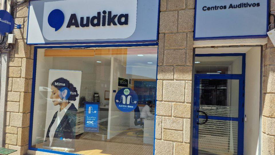Fachada de una clínica auditiva Audika, con rótulo ’Centros Auditivos’ y escaparate promocionando audífonos desde 49€. Interior visible con carteles y mobiliario especializado en cuidado auditivo.