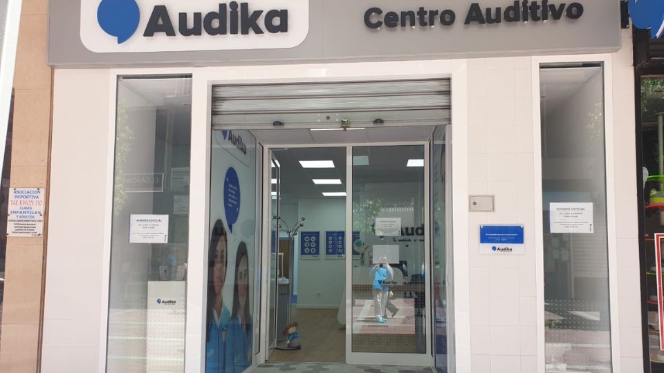 Fachada de un centro auditivo Audika con puertas de vidrio abiertas. En el interior, se observan carteles promocionales relacionados con audífonos y pruebas de audición. Letrero: 'Audika Centro Auditivo'.