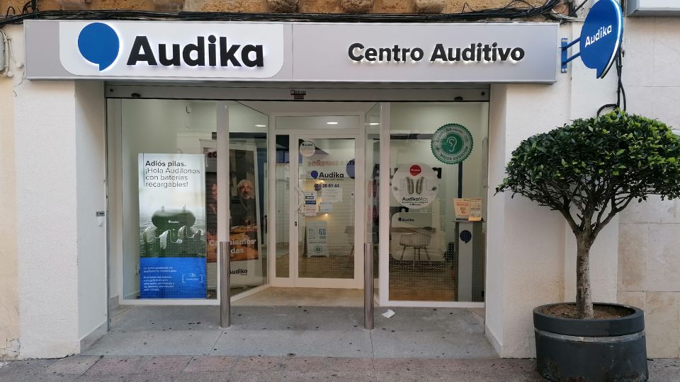 Fachada de un centro auditivo Audika con puertas de cristal; un cartel en la ventana dice: 'Adiós pilas. Hola Audífonos con baterías recargables.' Ambiente urbano con árbol decorativo.