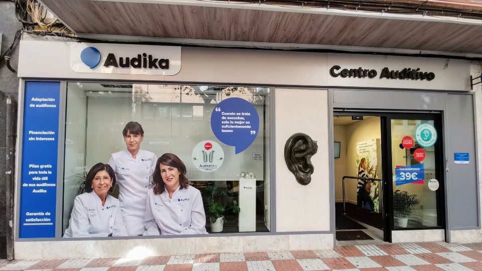 Fachada de un centro auditivo Audika, con cartel ’Centro Auditivo’. A la izquierda, vitrina con tres profesionales en bata blanca, textos: ’Adaptación de audífonos’, ’Financiación sin intereses’, ’Pilas gratis...’, ’Garantía de satisfacción’. Puerta derecha con publicidad: ’Tus audífonos desde 39€’. Ambiente urbano.