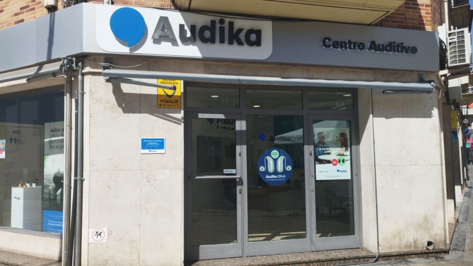 Fachada de un centro auditivo Audika con el logo azul sobre un rótulo que dice ’Audika Centro Auditivo’. Ventanas muestran publicidad de audífonos recargables. Cartel de seguridad: ’VISEGUR’.
