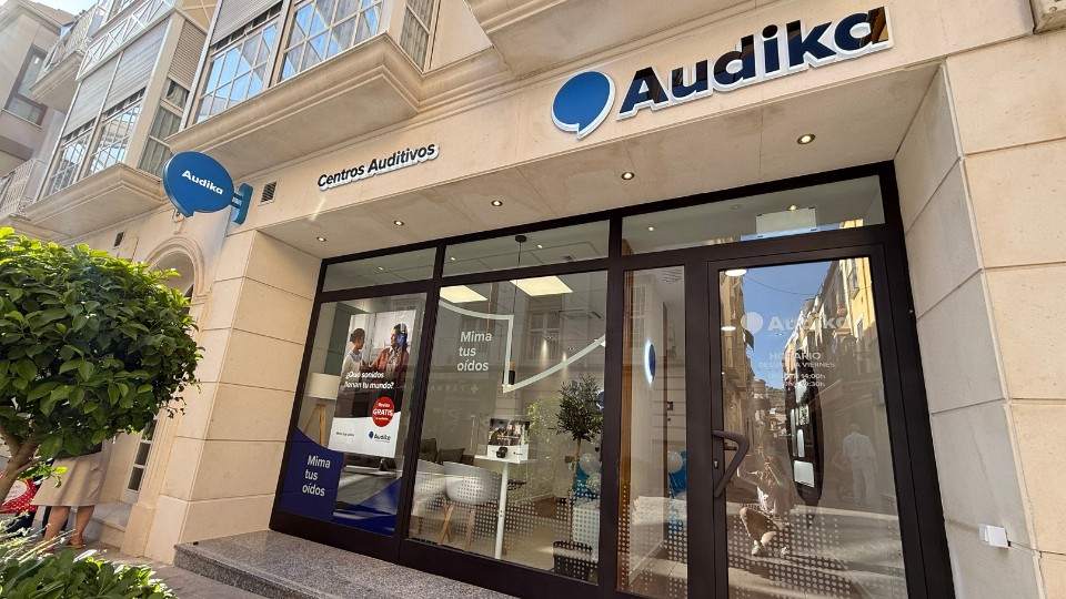 La fachada de un centro auditivo Audika muestra un letrero con ’Centros Auditivos’ y puertas de cristal. Un cartel promociona: “Mima tus oídos’. Ubicación urbana y luminosa.