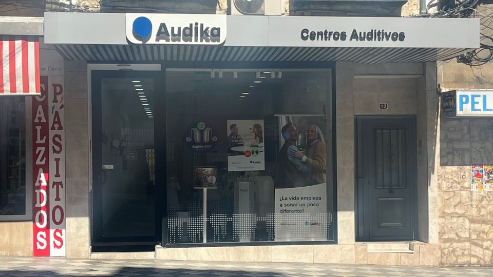 Fachada de un centro auditivo Audika con rótulo ’Centros Auditivos’. En el escaparate se observan carteles promocionales que incluyen el texto: ’La vida empieza a sonar un poco diferente’. Contexto comercial urbano con locales vecinos: una zapatería (’Calzados Pasitos’) y otra puerta sin identificación clara.
