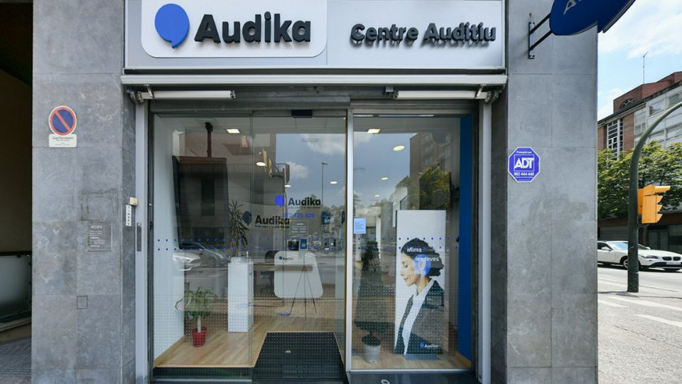 Fachada de una clínica auditiva Audika con rótulos ’Audika Centre Auditiu,’ puerta de vidrio, interior minimalista con muebles blancos, plantas y carteles promocionales. Texto visual: ’Milima, te atreves.’