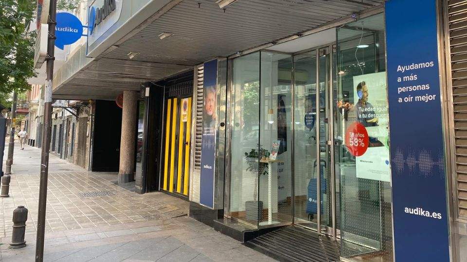 Fachada de una clínica Audika con un cartel que dice: 'Ayudamos a más personas a oír mejor', y texto adicional: 'audika.es'. Interior visible con carteles promocionales de audífonos.