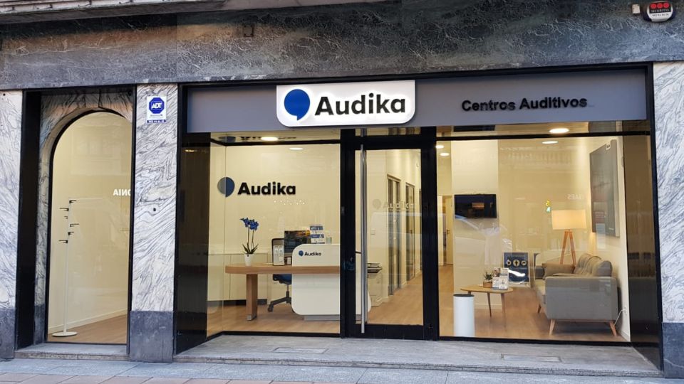 La fachada de una clínica auditiva de Audika muestra un rótulo iluminado con ’Audika’ y ’Centros Auditivos’. En el interior, destacan una recepción moderna, sofás y decoración minimalista.