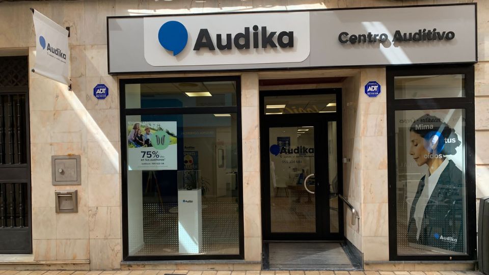 Fachada de un centro auditivo Audika con escaparates de vidrio. En el interior, se observa mobiliario de clínica. Cartel promocional destaca: '75% de descuento en audífonos recargables'.