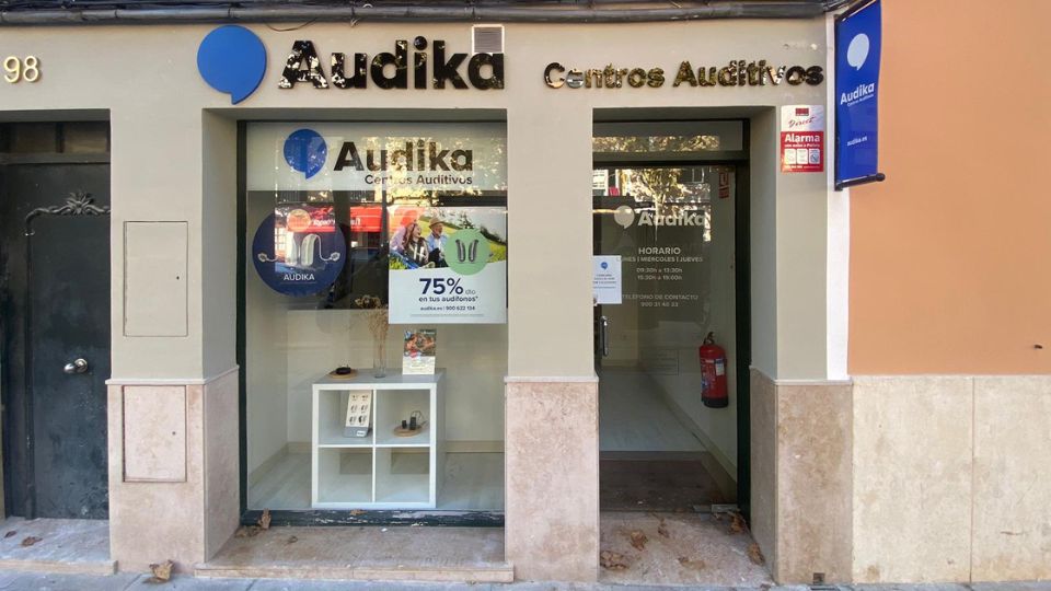 La fachada de un centro auditivo Audika muestra un escaparate con publicidad, incluyendo '75% en tus audífonos*'. Dentro, vitrinas exhiben audífonos. Contexto: calle urbana con acceso peatonal.