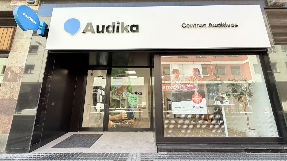 Fachada de una clínica auditiva Audika con letrero 'Centros Auditivos'. En el escaparate, se promocionan pruebas de audición gratis y el mensaje: '¿Qué sonidos llenan tu mundo?'. Contexto urbano.