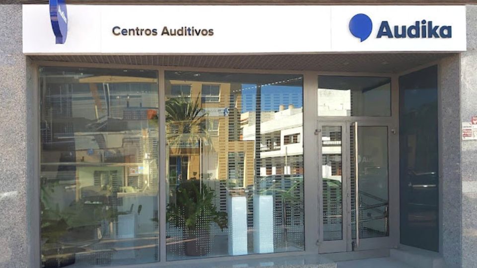 Fachada de un centro auditivo Audika, con ventanas de cristal que reflejan edificios y plantas en su interior. Letrero superior blanco con texto: 'Centros Auditivos' y el logo.