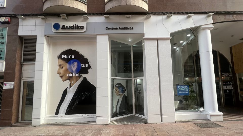 Fachada de un centro auditivo Audika con el letrero ’Mima tus oídos’ en una imagen publicitaria grande. En el cristal derecho destaca: ’PRUEBA GRATIS, CÓMODA Y FÁCIL’.