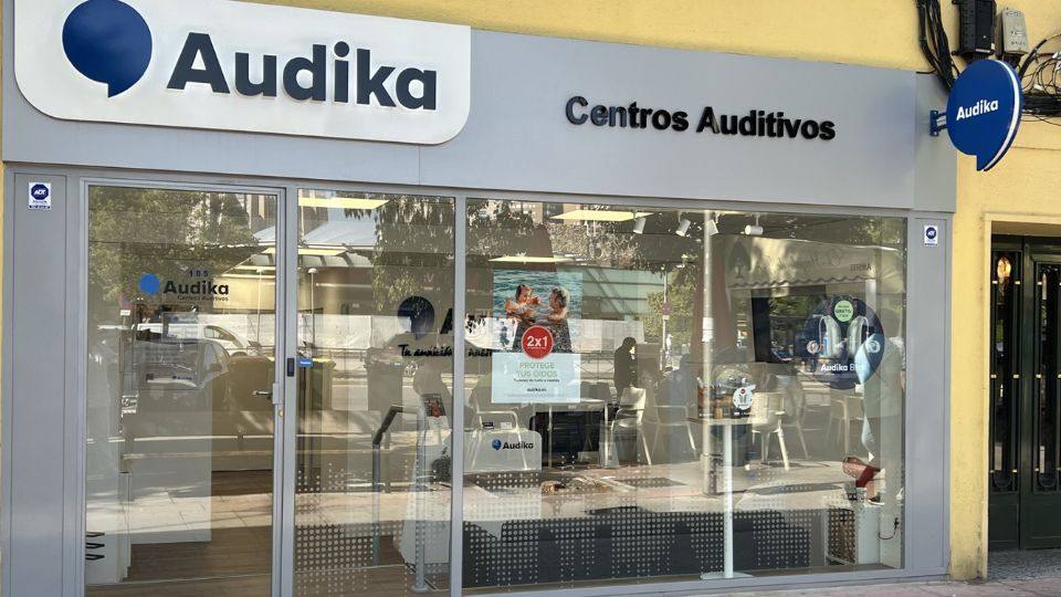 Fachada de una clínica auditiva Audika con cristales transparentes, el logo grande en la parte superior y rótulos que anuncian 'Centros Auditivos' y una oferta '2x1 Protege tus oídos'.