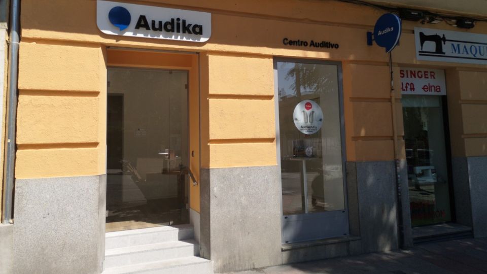 Fachada de un centro auditivo Audika con puerta de vidrio y ventana lateral. Letrero superior: ’Audika’ y ’Centro Auditivo’. Contexto: edificio amarillo, locales vecinos visibles.