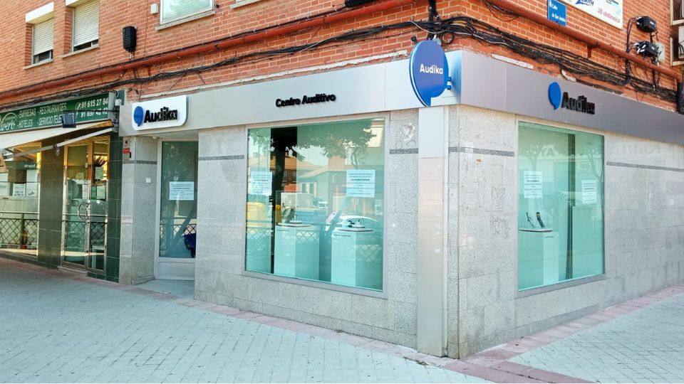 La fachada de un centro auditivo Audika muestra ventanas amplias con carteles informativos en sus vitrinas. El entorno es urbano, con edificios de ladrillo y aceras peatonales.