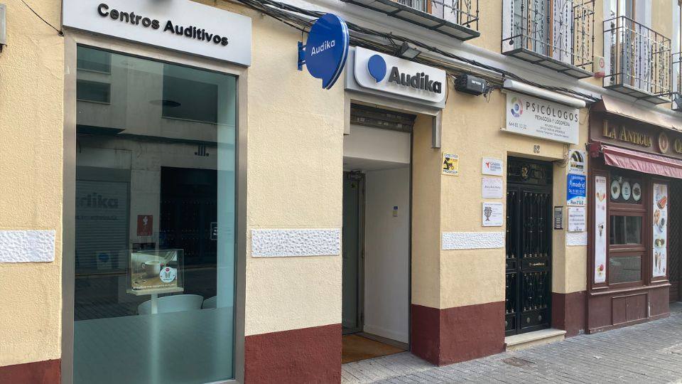 Fachada de una clínica de Audika con el letrero 'Centros Auditivos' y logotipo azul. A la derecha, otras tiendas y calles urbanas complementan el contexto. No se ven audífonos específicos.