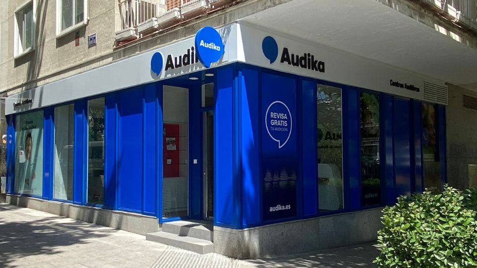 Fachada de un centro auditivo Audika, con diseño azul y blanco. Texto destacado: 'REVISA GRATIS TU AUDICIÓN', sitio web visible 'audika.es'. Contexto: calle urbana con vegetación cercana.