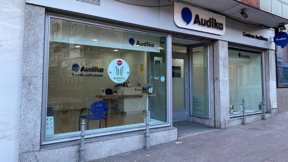 Fachada de un centro auditivo Audika con amplias ventanas que muestran un interior ordenado con sillas y un mostrador. El logo de Audika y el texto 'Tu oído es nuestra prioridad' destacan junto a un cartel promocionando audífonos. Ubicación urbana con acera frente al local.