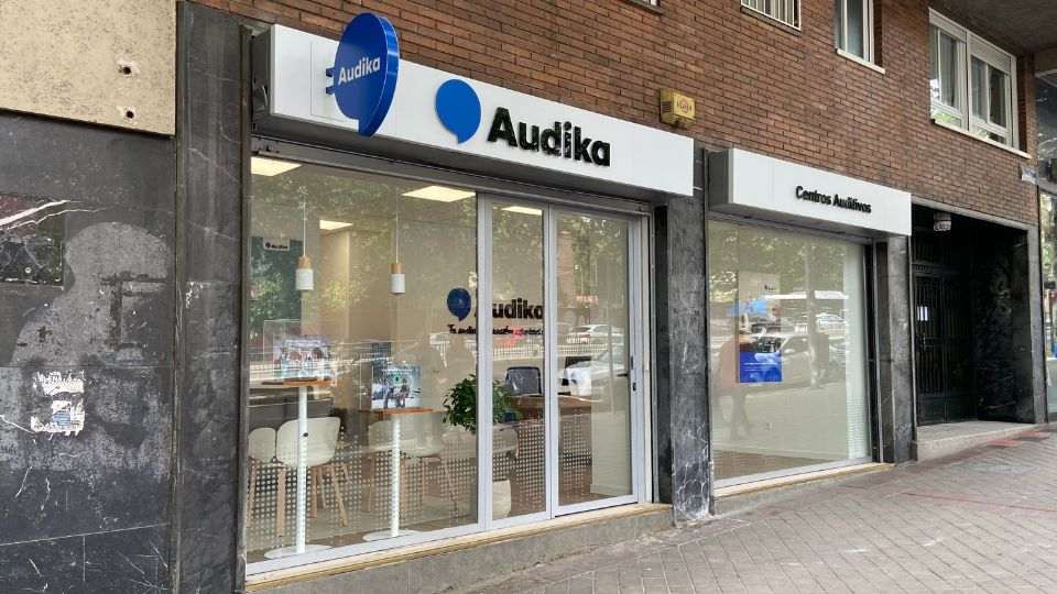 Fachada de una clínica auditiva Audika con grandes ventanas de cristal. Muestra el logo azul de Audika y el rótulo 'Centros Auditivos' en un edificio urbano de ladrillo.
