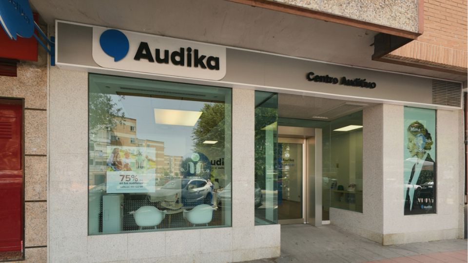 Fachada de un centro auditivo Audika con letrero ’Centro Auditivo’. Ventana muestra oferta: ’75% en tus audífonos’ junto a imágenes de audífonos y reflejo de edificios cercanos.
