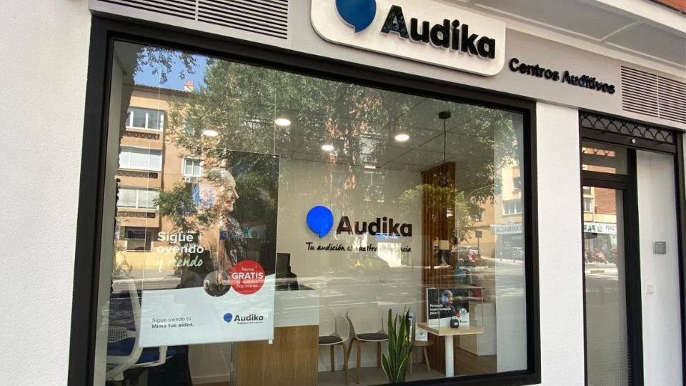 Fachada de una clínica auditiva Audika con letrero 'Centros Auditivos'. En la vitrina, carteles destacan 'Sigue oyendo y viviendo' y 'Revisión GRATIS hoy mismo'. Interior minimalista con sillas y planta.
