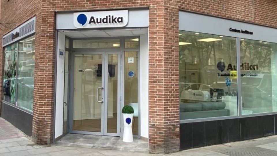 La fachada de una clínica Audika, especializada en cuidado auditivo, muestra un letrero con el logo azul. Al interior, visible a través de ventanales, destacan muebles modernos.