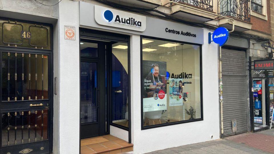 Fachada de un centro auditivo Audika con letreros que dicen 'Centros Auditivos' y 'Sigue oyendo y haciendo cosas gratis'; ventanas grandes muestran carteles promocionales. Ubicado en una calle urbana.