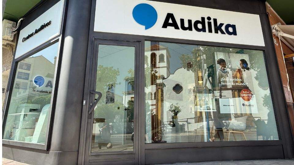 Fachada de un centro auditivo Audika, con grandes ventanas de cristal que reflejan el exterior. Texto visible: “¿Qué sonidos llenan tu mundo?’, “Prueba GRATIS’, y el logo de Audika.