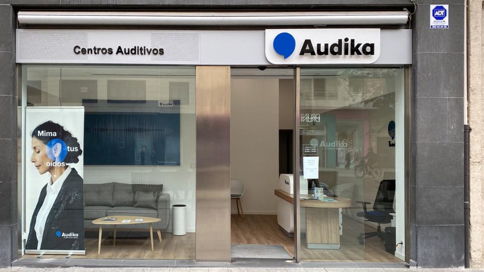 Fachada de un centro Audika con puertas de vidrio abiertas. Texto visible: 'Centros Auditivos' y 'Mima tus oídos'. Interior moderno con sofá, mesa, escritorio y silla; especializado en audífonos.