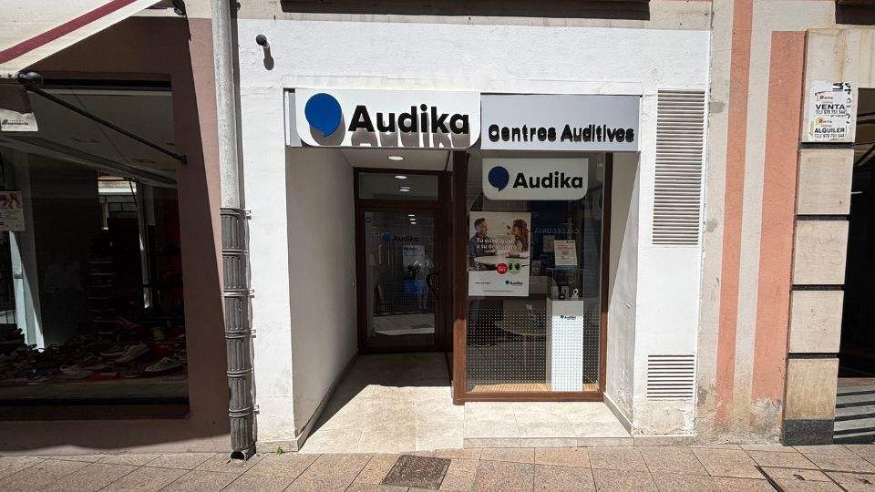 Fachada de una clínica auditiva Audika con el texto ’Centres Auditius’ en el cartel superior. En el escaparate hay pósters promocionales y referencias a audífonos. Calle peatonal urbana alrededor.