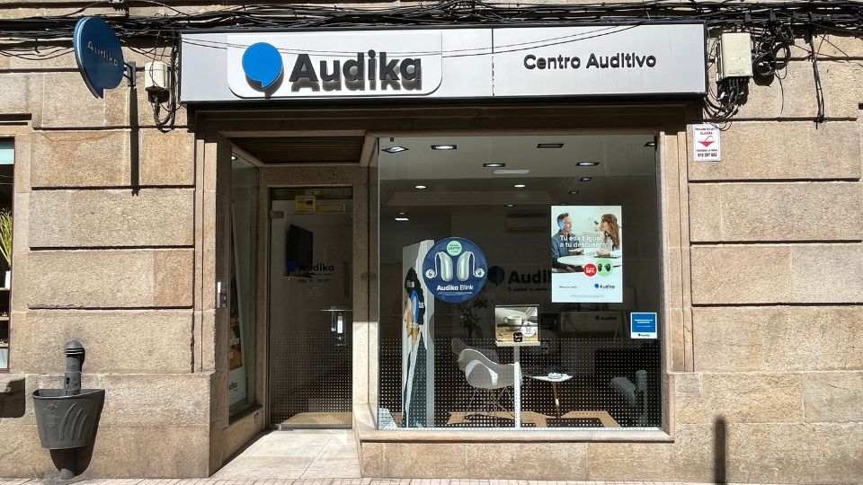 La fachada de un **centro auditivo Audika** muestra un letrero con el logo y texto ’Audika Centro Auditivo.’ A través del escaparate, se observan expositores de audífonos, como el modelo **Audika Blink**. En el interior, hay carteles promocionales, uno indicando: ’Tú das el primer paso, tu oído te lo agradecerá. 15%.’ El entorno es una calle urbana con paredes de piedra y una papelera cercana.