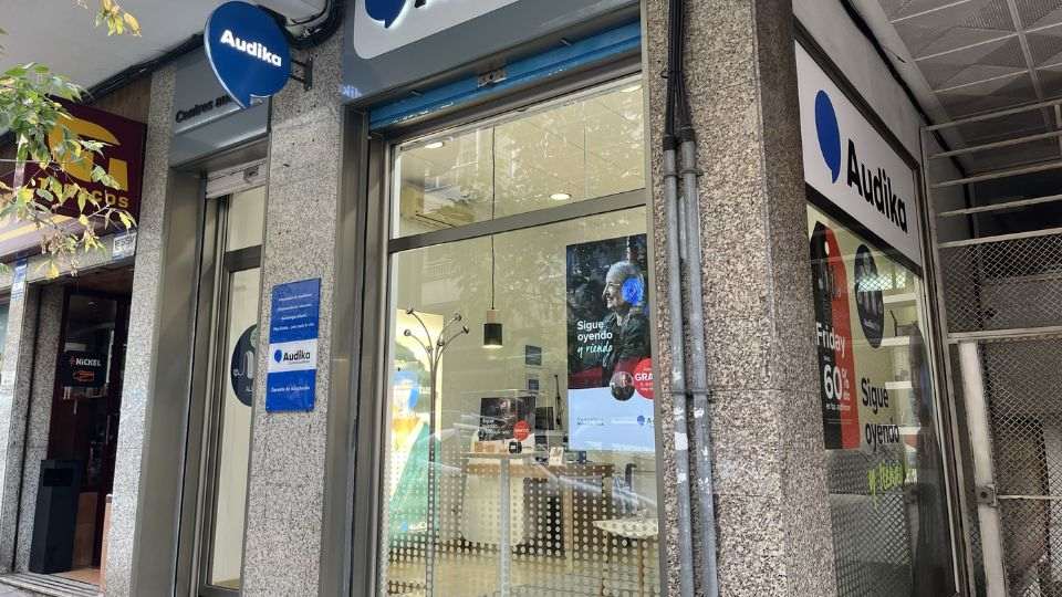 Fachada de una clínica auditiva Audika con logotipo azul visible; vitrinas muestran carteles promocionales: 'Sigue oyendo el mundo,' 'GRATIS prueba auditiva.' Rodeada por negocios urbanos, interior iluminado.