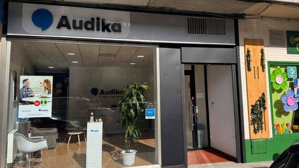 Fachada de una clínica Audika especializada en cuidado auditivo, con gran cristalera mostrando el interior moderno. Un póster anuncia: 'Tu edad juega a tu descuento', destacando audífonos Audika.