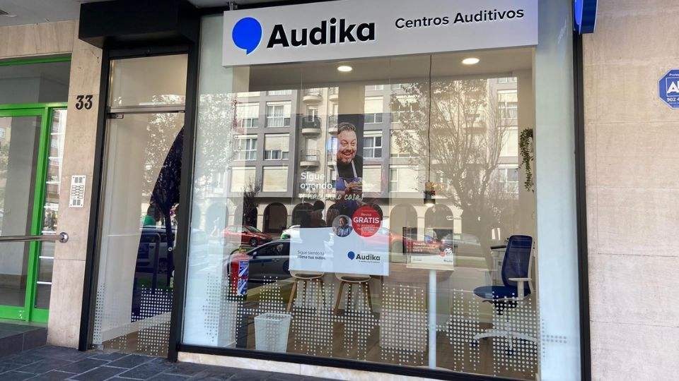 Fachada de una clínica Audika, especializada en cuidado auditivo, con ventanas que muestran un interior moderno. Texto visible: ’Audika Centros Auditivos’, ’Sigue oyendo lo que importa’, ’Prueba gratis’. Dirección: número 33.