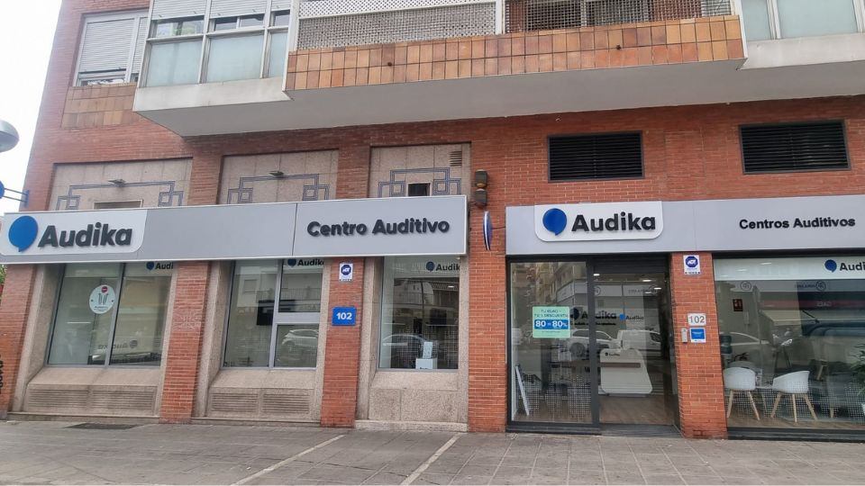 Fachada de un centro auditivo Audika con rótulos que dicen 'Centro Auditivo' y 'Centros Auditivos', ubicado en un edificio ladrillo. Dirección visible: 102. Horario en puerta: '10-18h'.